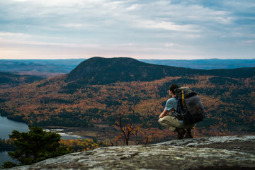 a man with a backpack kneels on top of a mountain in maine in the fall with autumn foliage and a lak