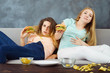 Overeating, sedentary lifestyle, fast-food, laziness, pleasure, delight, enjoyment, appetite, hunger. Two young lazy women lying at coach eating burgers and chips