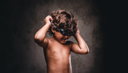 Cute little shirtless boy puts on swimming goggles posing in a studio. Isolated on the dark textured background.