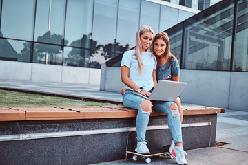 Wall Mural - Two beautiful hipster girls sitting on the bench with a laptop on a background of the skyscraper.