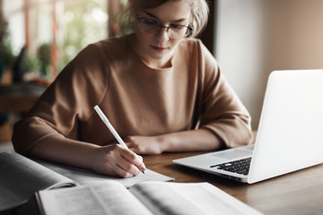 Close-up shot of hardworking attractive female entrepreneur in glasses, writing in notebook, checking spelling in dictionary, making homework for university, using laptop to browse in network