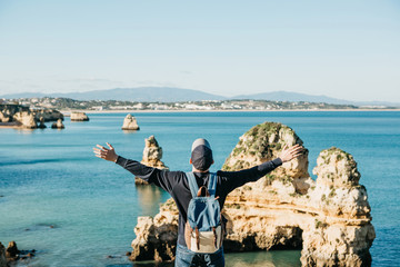 Wall Mural - A tourist or traveler with a backpack admires the beautiful view of the Atlantic Ocean and the coast near the city called Lagos in Portugal and raises his hands up showing how he is free and he likes.