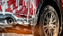 Red Compact SUV Car With Sport And Modern Design Washing With Soap. Car Covered With White Foam. Car Care Service Business Concept. Car Wash With Foam Before Glass Waxing And Glass Coating Automobile.