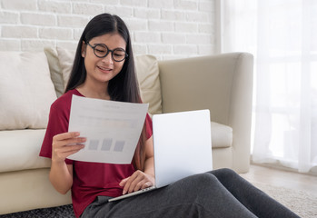 Poster - asian woman freelance wear eyeglasses working on laptop with paperwork sitting one floor and leaning sofa at home,work at home concept.