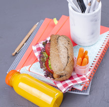 A Sandwich With Sausages And Salad In A Lunch Box With Vegetables And Fresh Juice In A Bottle. Delicious And Healthy Lunch For School And Office. Books And Pencils For Study