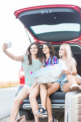 Sticker - Three young women with suitcases on a trip by car. They sit in the back of the car, they look at the map, resting after a long drive and having fun. Hitchhiking and car trips with friends