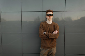 Wall Mural - Handsome young man with vintage sunglasses in a fashionable sweater stands near the wall