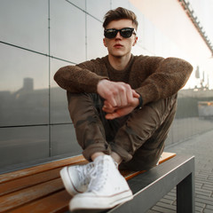 Wall Mural - Fashionable stylish man model in sunglasses in a pullover and fashionable military pants with white sneakers sits on a bench