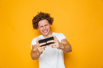 Poster - Happy gamer man in casual t-shirt playing online video games on smartphone, isolated over yellow background