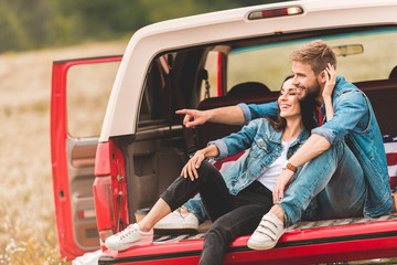 Wall Mural - beautiful young couple relaxing in car trunk during trip and pointing somewhere