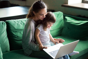 Happy mother with little kid daughter having fun online with application, watching video cartoons or playing game on laptop together, young babysitter or nanny teaching child girl to use computer