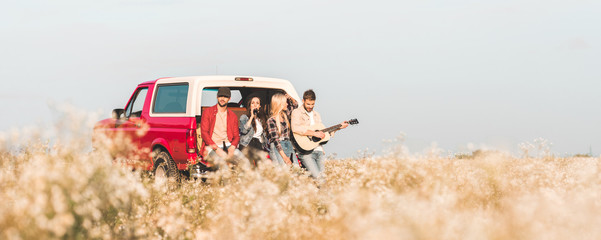 Wall Mural - group of young friends drinking beer and playing guitar while relaxing in car trunk in flower field