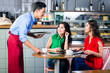 © Kzenon - Side view of a handsome and polite waiter smiling while serving coffee at the table of two beautiful young women in a trendy coffee shop