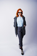 Portrait of a fantastic redheaded girl in blue shirt, grey overcoat posing with sunglasses in the studio.