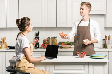 Wall Mural - girlfriend using laptop, boyfriend holding bottles of juice in kitchen