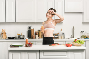 Wall Mural - attractive girl in sport bra touching forehead and holding bottle of fresh juice in kitchen