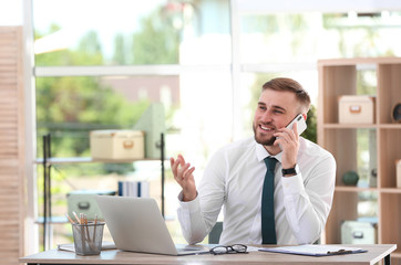 Poster - Young businessman talking on phone while using laptop at table in office