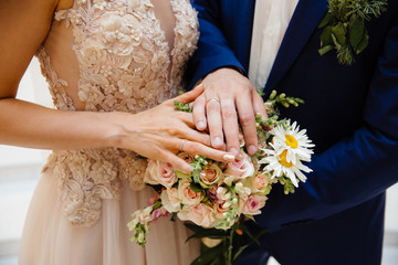 A close-up of a cropped frame of the bride and groom standing side by side and touching their wedding bouquet, showing their wedding rings on the pawls.