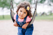 © anoushkatoronto - Portrait of happy smiling little latin hispanic toddler girl swinging on swings at playground outside on summer day. Happy childhood lifestyle concept. Toned with film pastel faded filters colors.