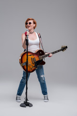 Wall Mural - Young woman musician with an acoustic guitar in hand on a gray background. He laughs and plays rock and roll loudly. Full-length portrait