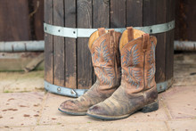 Old Cowboy's Boots Without Spurs Stand On The Street Near A Wooden Brown Barrel In The Afternoon.
