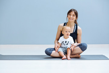 Wall Mural - Happy sportive brunette mother hugging and playing with her lovely bay toddler son while sitting on floor over grey background. Fitness, happy maternity yoga with children concept.