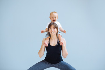 Cute european sporty mother playing with cute toddler baby boy. Little son sitting on his mother's shoulders at cozy simple home interior. Motherhood, healthy lifestyle and people concept.