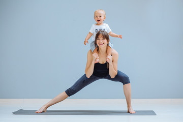 European sportive mother piggybacking her toddler baby son in fitness clothing on gray background. Lounge exercise. Motherhood, healthy lifestyle concept.