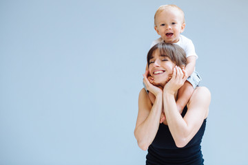 happy blond toddler boy laughing on to his sportive mother for a piggyback ride over gray background
