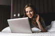 © Westend61 - Smiling woman lying on bed at home looking at laptop