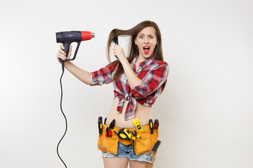 Silly mad strange fun handyman woman in plaid shirt, denim shorts, kit tools belt full of instruments dry hair with power heat gun isolated on white background. Female in male work. Renovation concept