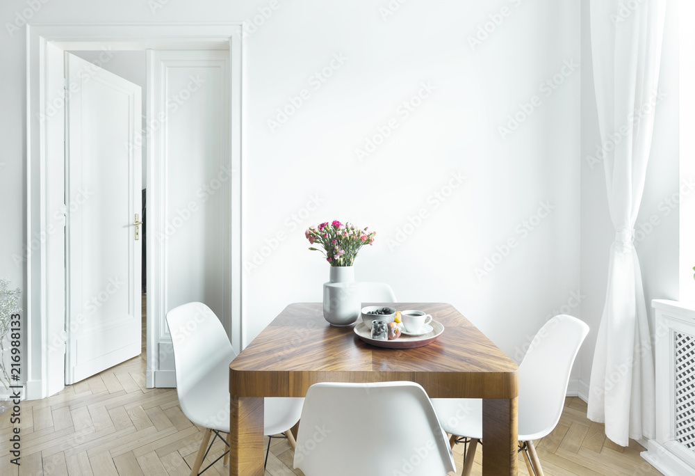 Dining table with fresh flowers and breakfast tray with coffee cup and fruits in real photo of white room interior with empty wall. Paste your painting here - obrazy, fototapety, plakaty 