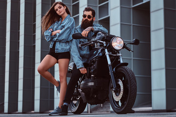 Attractive hipster couple - bearded brutal male in sunglasses and jeans jacket sitting on a retro motorcycle and his young sensual girl standing near, posing against a skyscraper.