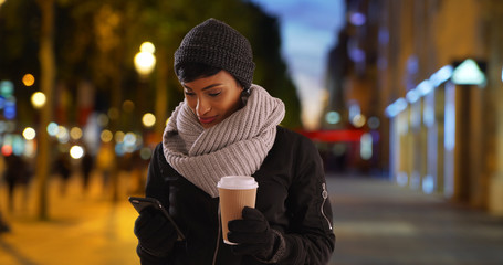 Wall Mural - Pretty black woman with warm cup of coffee using smartphone outside