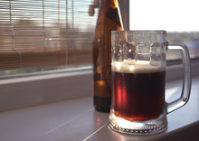 A Large Glass Mug With A Dark Beer And A Bottle Of Brown Glass Is On The Windowsill. The Sun Passes Through The Drink.