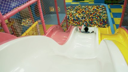 Wall Mural - Rear view of little black girl of elementary school age sliding down into ball pit when having fun on plastic slides in playroom