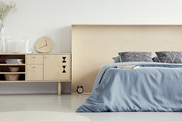 Wall Mural - Real photo of wooden cupboard with glass vases, minimal clock and books standing in white bedroom interior with bed with pastel blue sheets in box headboard