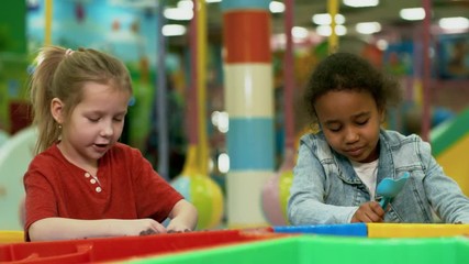 Wall Mural - Medium shot of two little girls of different ethnicities playing with sand molds and shovels in sandbox of indoor amusement park