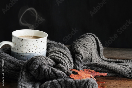 Fotografie, Obraz Hot steaming cup of coffee with cream wrapped in a cozy grey scarf with fallen leaves for autumn