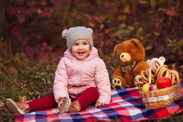 Portrait of beautiful happy child sitting on plaid with autumn colorful leaves background. Picnic basket with apples and bagels, toy bears. Funny girl outdoors in fall park.