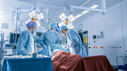 Shot of Diverse Team of Professional Surgeons Performing Invasive Surgery on a Patient in the Hospital Operating Room. Anesthesiologist Monitors Vitals. Modern Hospital.