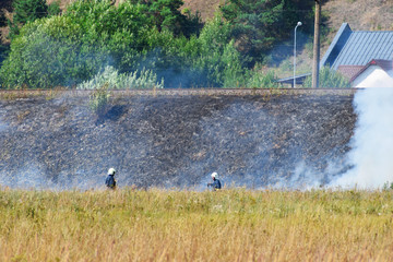 Wild fire and smoke in dry meadow grass due to hot windy weather in summer started to burn from railway behind and two firefighters wearing  helmets working. Firefighting rescue operation concept.