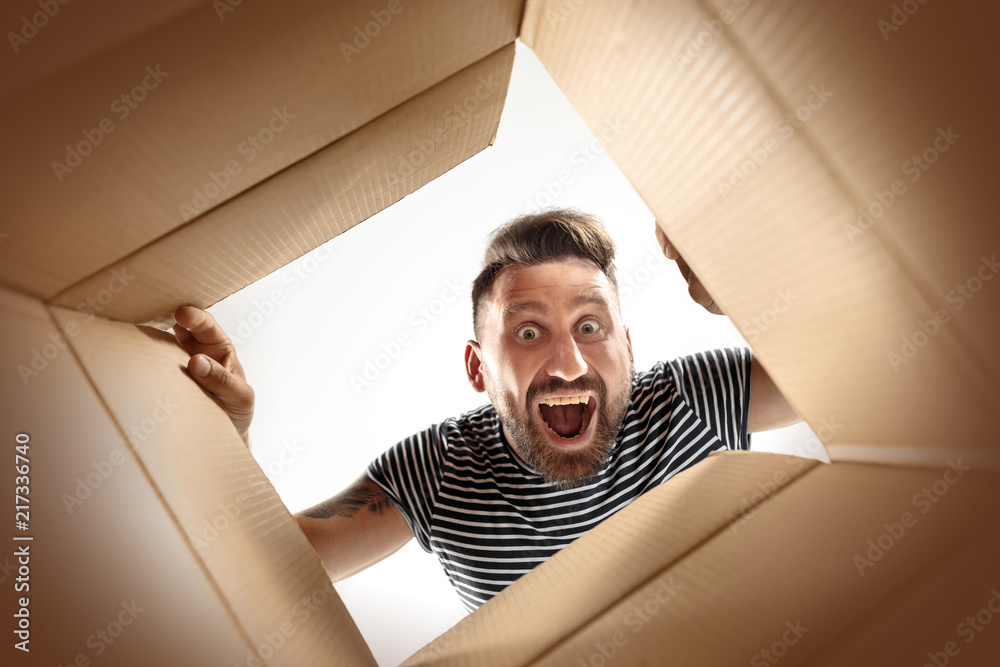The surprised man unpacking, opening carton box and looking inside. The package, delivery, surprise, gift lifestyle concept. Human emotions and facial expressions concepts - obrazy, fototapety, plakaty 
