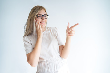 Smiling cheerful young woman in glasses presenting new information. Beautiful Caucasian girl pointing finger at copy space and touching cheek in admiration. Promotion concept
