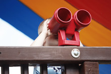 Little Blond Boy Looks Through Binoculars On A Playground.