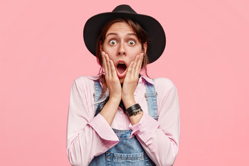 Wall Mural - Photo of surprised female gardener opens mouth widely, keeps both hands on cheeks, reacts on something terrible, dressed in shirt, overalls, black hat, isolated over pink background. Omg, I am shocked