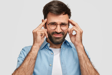 Desperate unshaven male keeps hands on temples, frowns face in displeasure, suffers from headache, dressed casually isolated over white background. Handsome man expresses frustration, negative feeling