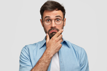 Photo of surprised bearded male holds chin and looks in hesitation, wonders latest news, wears spectacles and elegant blue shirt, isolated over white background. People and facial expressions concept