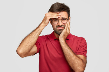 Studio shot of dissatisfied bearded male has toothache and headache, feels unhappy and tired after long work, frowns face in discontent, dressed in red t shirt, isolated over studio background