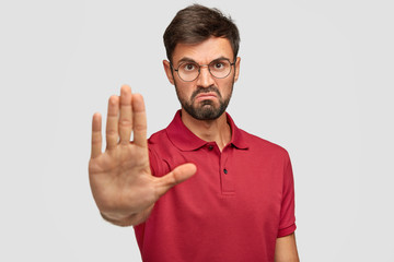 It`s forbidden! Angry displeased young male frowns face, shows stop gesture, keeps palm in front, tries to prevent himeself from something bad and unpleasant, wears casual t shirt, isolated on white